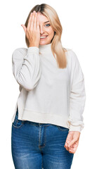 Young caucasian woman wearing casual winter sweater covering one eye with hand, confident smile on face and surprise emotion.