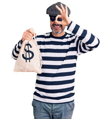 Middle age handsome man wearing burglar mask holding money bag smiling happy doing ok sign with...