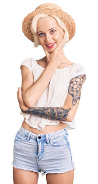 Young blonde woman with tattoo wearing summer hat looking confident at the camera smiling with crossed arms and hand raised on chin. thinking positive.