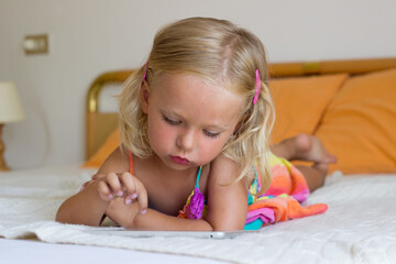 Little blond girl lay on bed and play tablet,toddler using internet with gadget,kid addicted to tablet and looking cartoons,3 year old child studiyng online with developmental games