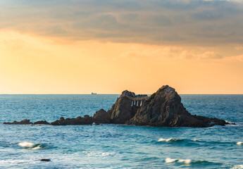 Seascape depicting a close up on the Sakurai Futamigaura's Couple Stones protected by a sacred shimenawa hemp or straw rope surrounded by a turquoise blue water with breaking waves at sunset.