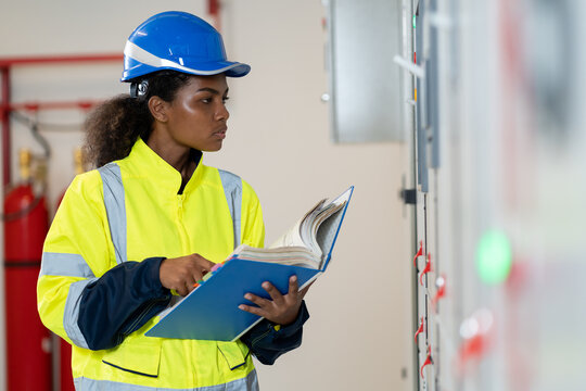 Female electrician engineer maintenance electric system with manual book in control room. African American electrician female engineer checking electric system in control room