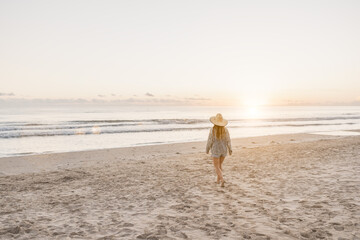 Blonde girl, wearing a hat, walking on the beach at sunrise.