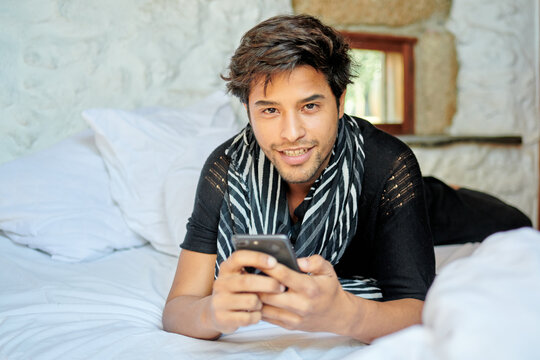 Stylish Hispanic man chatting on smartphone on bed