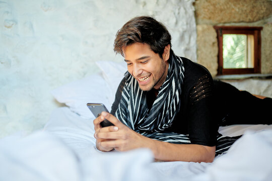 Stylish Hispanic man chatting on smartphone on bed