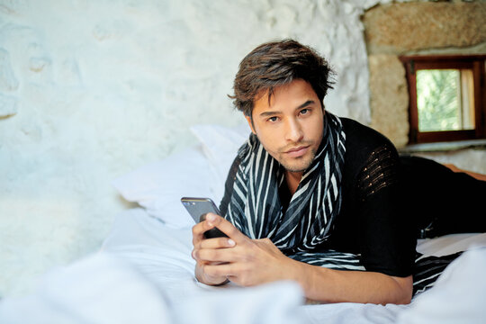 Stylish Hispanic man chatting on smartphone on bed
