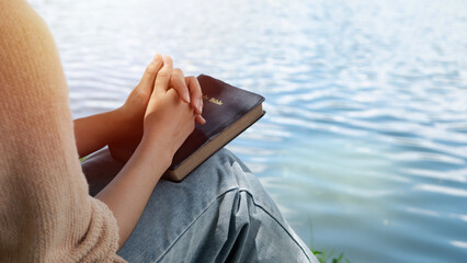 A woman puts her hands on the bible for devotion and prays alone at the lake in the morning during...