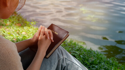 A woman puts her hands on the bible for devotion and prays alone at the lake in the morning during the sun rises. Christian background with copy space.