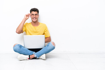 Young man sitting on the floor isolated on white background with glasses and happy