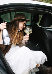 The bride in a white dress, leather jacket and hat sits in the back seat of the car, holding a bottle of champagne, a glass of champagne and smiling.
