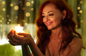 Happy beautiful woman looking at burning candles and making wish on Christmas eve. Portrait of a girl on bokeh background. New Year