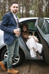The groom is standing near the gray car and holding a bottle of champagne. The bride is sitting in the back seat of the car, holding a glass of champagne and smiling.
