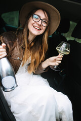 The bride in a white dress, leather jacket and hat is sitting in the back seat of the car, holding a bottle of champagne, a glass of champagne and laughing.