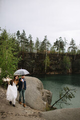 The bride and groom walk in the rain on a rock by the lake. The bride in a hat and leather jacket holds a wedding bouquet. The groom is holding a transparent umbrella.