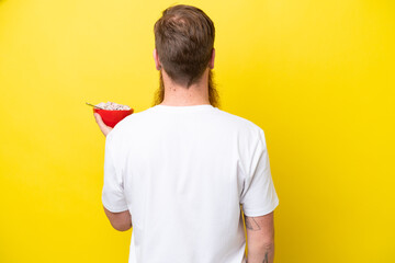 Redhead man with beard eating a bowl of cereals isolated on yellow background in back position