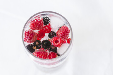 Carbonated drinks with fresh berries and ice. Fresh cold sparkling bubble water with raspberry, black and red currant berries in transparent glass with ice, top view macro. Food and drink concept