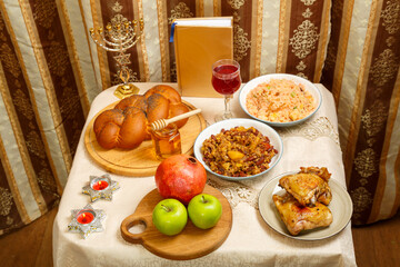 Festive table for a meal on Rosh Hashanah challah, honey, pomegranate, apples, wine and traditional dishes next to the menorah and siddur.