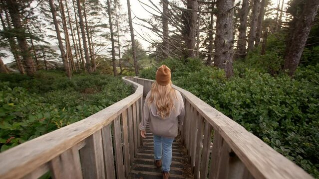 
Young women walks down picturesque boardwalk surrounded by green bushes and tall trees, canadian west coast
