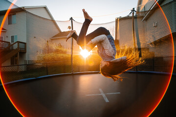 A girl on a trampoline is suspended mid-flip at sunset with a sunflare