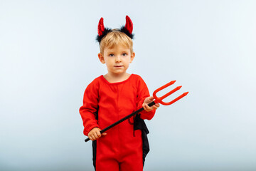 Happy Halloween! Cute little boy in devil halloween costume with horns and trident on light blue...
