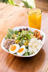Tuna salad, egg, potato, lettuce, onion, tomato, corn, black olives and asparagus. Served in a white bowl on a wooden table. Healthy meal.