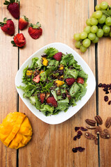 Salad of lettuce, mango, strawberry, nuts and seeds. Served in a white bowl on a wooden table. Healthy meal.