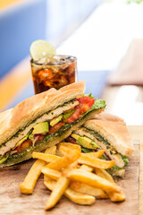 Chicken sandwich, avocado, red onion, tomato, lettuce and pesto, served with french fries and a glass of soda. Served on a wooden board