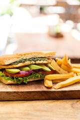 Chicken sandwich, avocado, red onion, tomato, lettuce and pesto, served with french fries. Served on a wooden board in a restaurant.