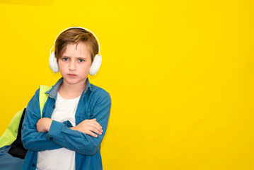 serious dissatisfied schoolboy listens to music in headphones, stands with a backpack. yellow...