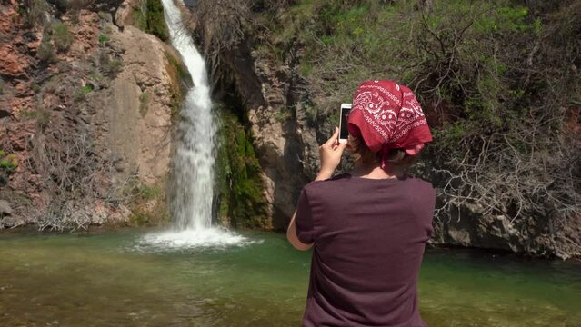 Tourist woman in red bandana takes photos of waterfall during hike, using mobile phone, view from the back. Young female blogger makes content for her travel vlog.