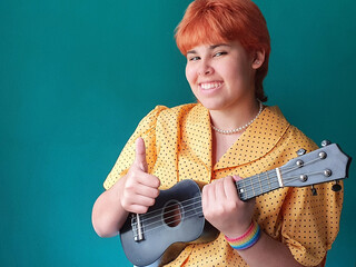 Happy queer female teenager portrait wearing a rainbow bracelet with a thumb up and holding ukelele instrument. LGBTQ people. Copy space.