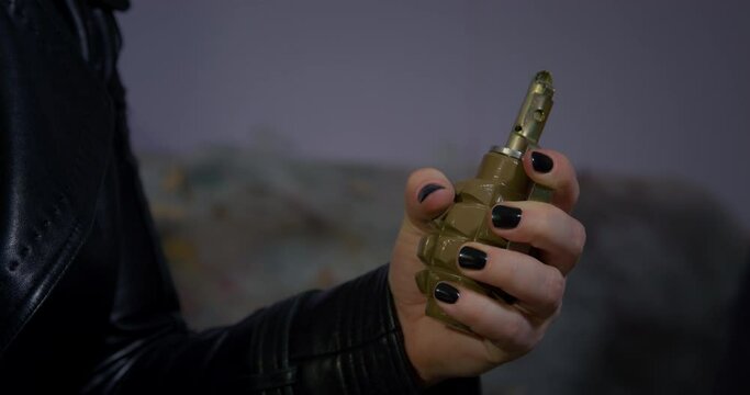 Criminal woman with black nails takes hand grenade out of leather jacket. Female killer activates dangerous projectile on blurred background close view