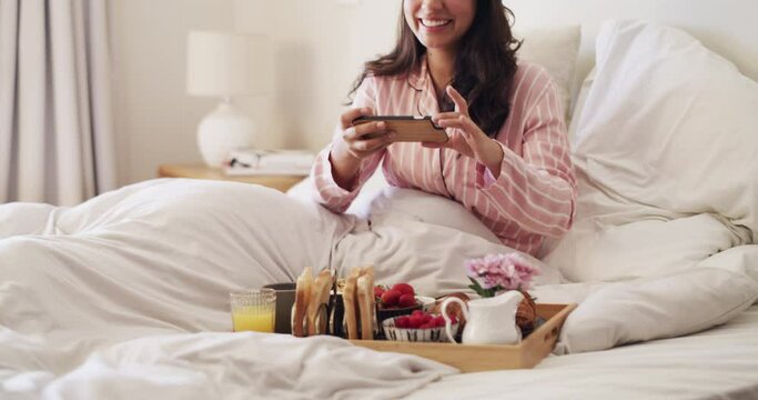 Female influencer taking a picture of breakfast in bed with a phone in her modern bedroom at home. Happy woman blogger in pajamas posting a healthy brunch on social media in a hotel room.