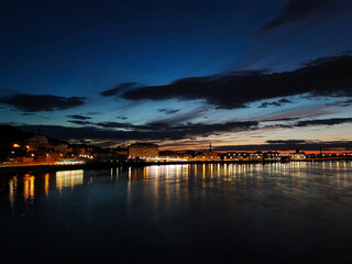 Night promenade in the distance by the river with illuminated houses. On the background of a dark blue sky with clouds