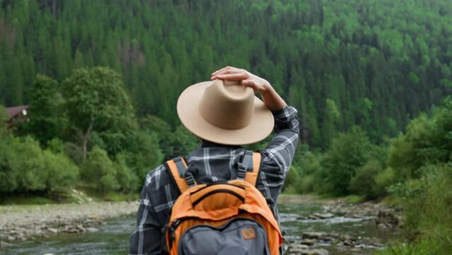 A tourist girl puts on a hat standing near the river, a mountain river a tourist puts on a hat against the backdrop of mountains. A traveler with a backpack stands against the backdrop of mountains
