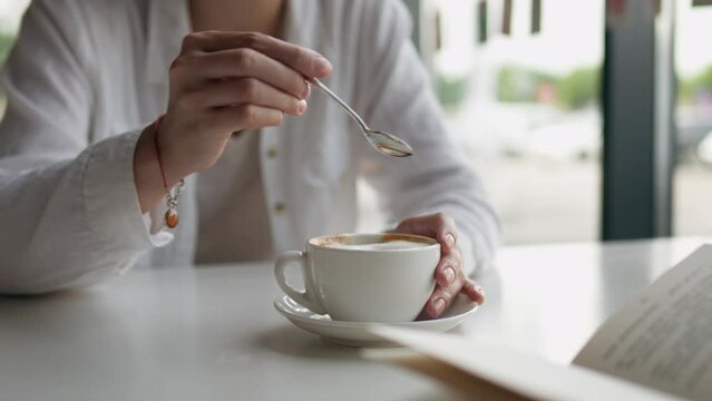 A woman sits in a cafe in front of large windows, drinks a cappuccino with thick foam, reads a book. Girl reading a book stirs cappuccino with a metal spoon
