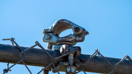 Metal clamp bolted to top of chain link fence with blue sky. 