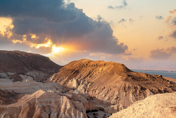 Obraz premium Evening gold sunset sky over mountain Sodom Gomorrah from Negev desert, Israel, Dead sea. Sunset on a large salt formation mountains range Sodom with fluffy clouds. Sun shining on rocky landscape.