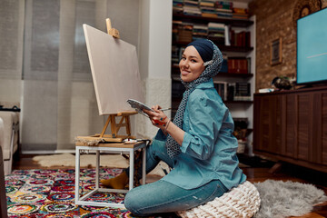 A woman in a hijab paints on canvas with a brush and tempera