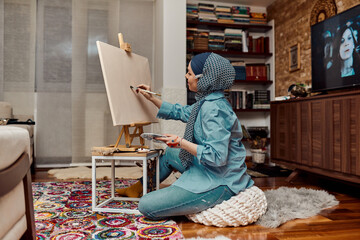 A woman in a hijab paints on canvas with a brush and tempera