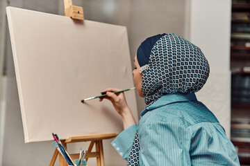 A woman in a hijab paints on canvas with a brush and tempera