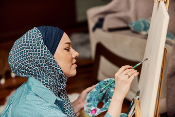 A woman in a hijab paints on canvas with a brush and tempera
