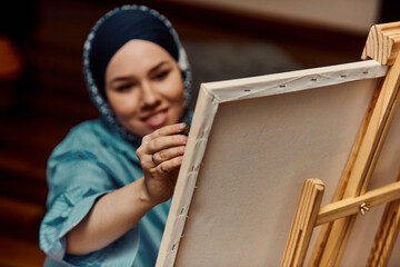 A woman in a hijab paints on canvas with a brush and tempera