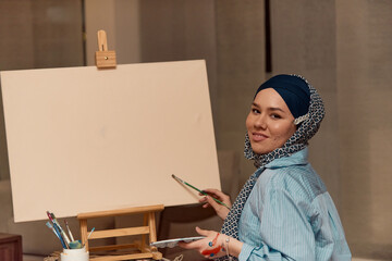 A woman in a hijab paints on canvas with a brush and tempera