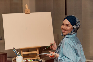 A woman in a hijab paints on canvas with a brush and tempera