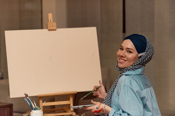 A woman in a hijab paints on canvas with a brush and tempera