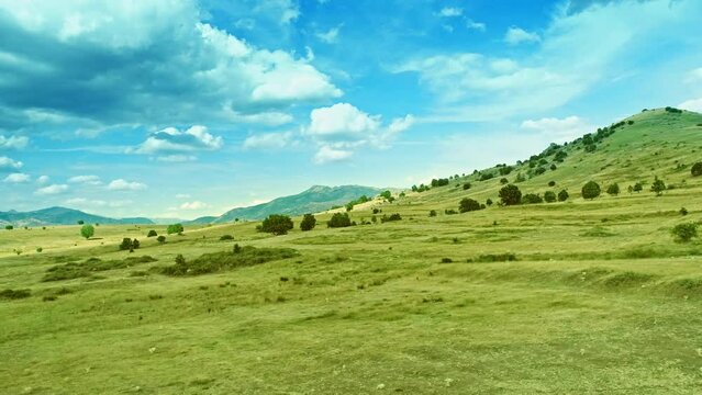 Lateral drone motion view along gentle slope, uneven grass covered terrain and cloudy sky