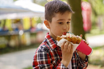Small Caucasian boy eating sweet waffle at street. Child with fast food in hands.