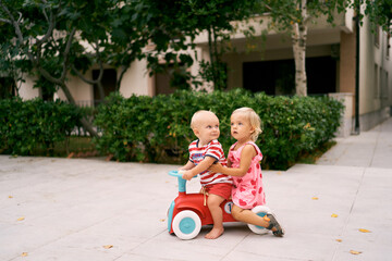 Little boy and girl are sitting on a toy car on a paved path. High quality photo