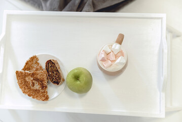 light photo with breakfast, coffee with marshmallows, green apple and pancakes on a white tray, top view, selective focus